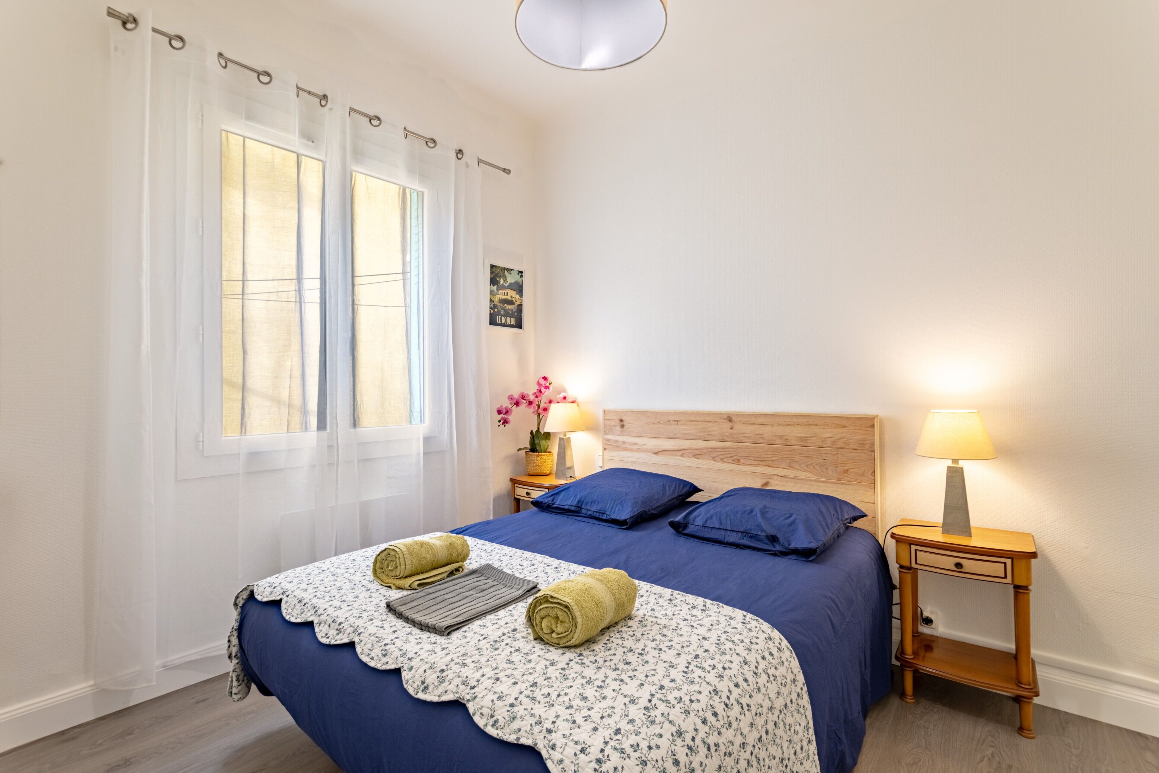 A bedroom with a bed, a nightstand, a lamp, a window with curtains, and a wall-mounted picture.