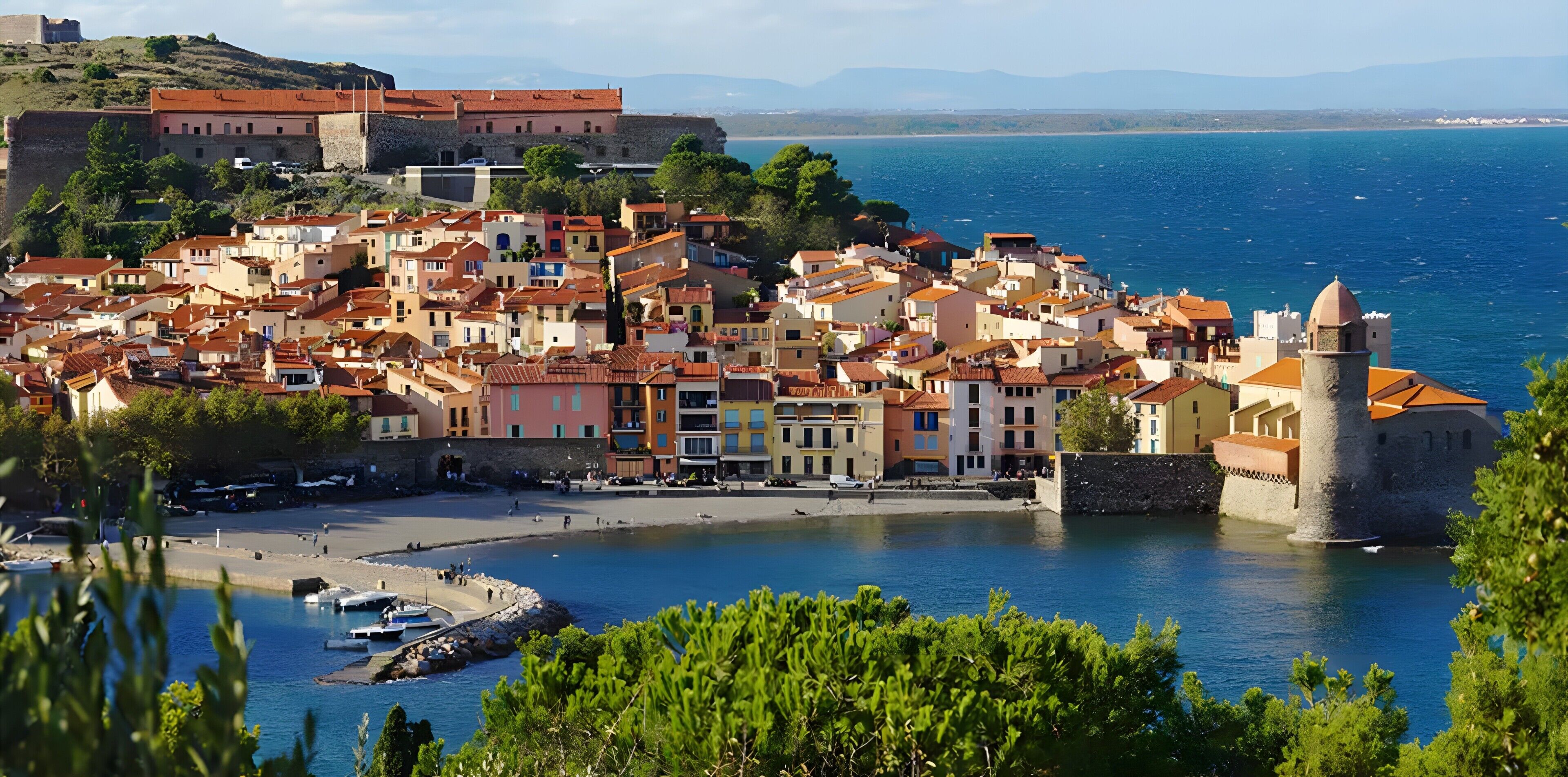 Uma vila costeira com edifícios históricos, uma estrutura fortificada e uma vista clara do mar.