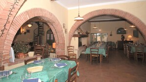 Un comedor con arcos de ladrillo, mesas y sillas de madera. Se observan varias mesas dispuestas con platos, vasos y cubiertos. La sala tiene un encanto rústico, con vigas de madera a la vista y una chimenea de ladrillo.