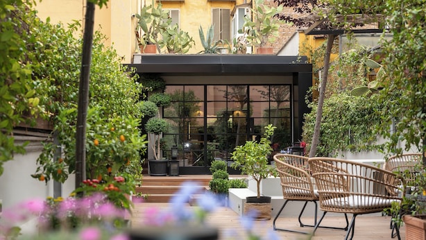 Un patio con sillas de mimbre, plantas en macetas y vistas a un edificio con grandes ventanales.