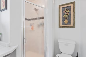 Kylpyhuone, jossa on valkoinen wc-istuin, lasinen suihkuovi ja seinällä kehystetty taideteos.