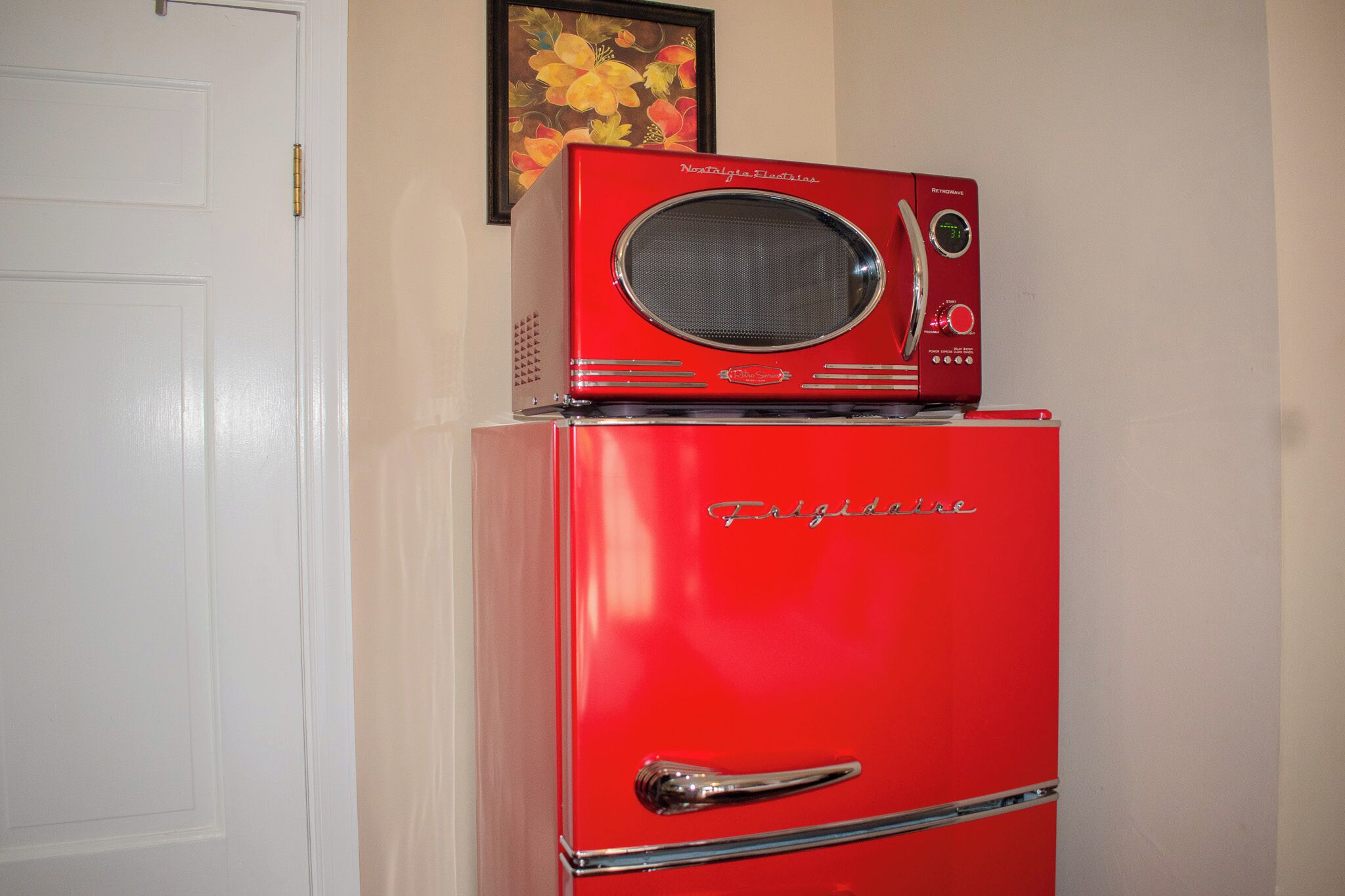Un vieux réfrigérateur rouge, de style rétro, avec un four à micro-ondes intégré sur le dessus, placé contre un mur, au-dessus duquel on aperçoit un tableau encadré.
