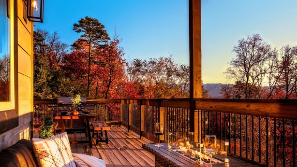 Una terraza de madera con mobiliario exterior, una mesa con vasos y un brasero, con vistas a árboles otoñales.