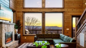 Un interior de cabaña acogedor con chimenea, ventanas amplias, un sofá modular y una escalera.