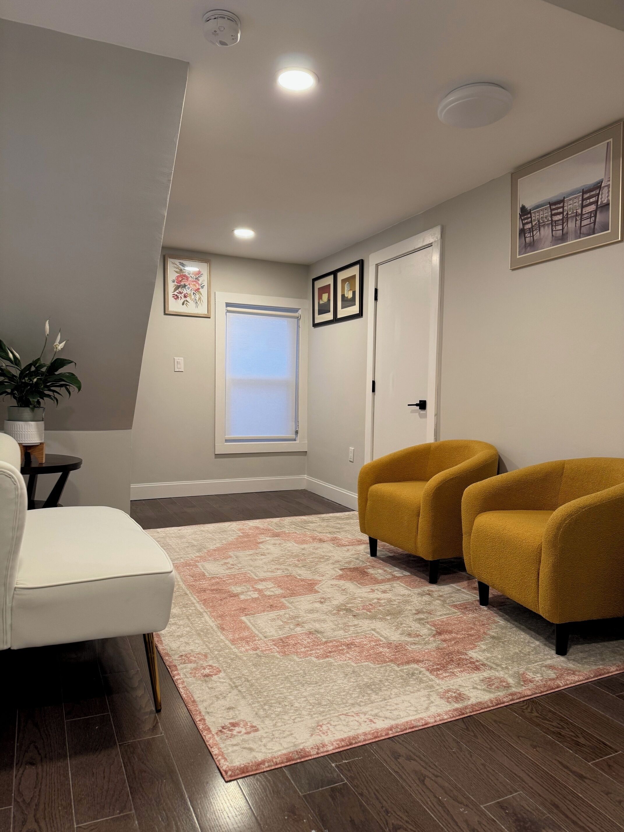 Een moderne woonkamer met een witte bank, twee mosterdgele fauteuils, een kamerkleed met dessin, en een groot raam met jaloezieën.