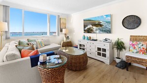 Una sala de estar con sofá, mesa de centro y mesita auxiliar, un armario blanco y un ventanal con vista al mar.