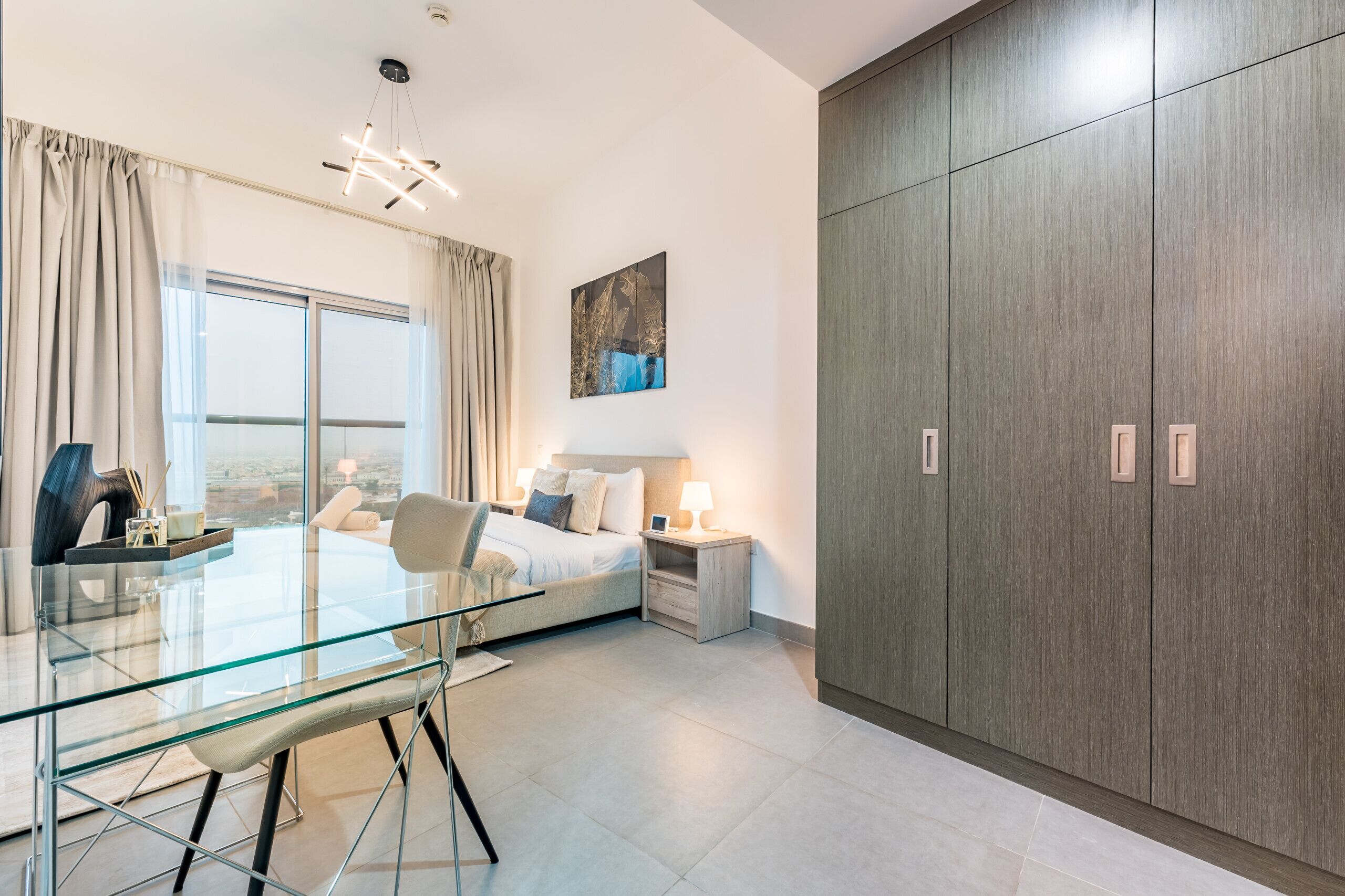 A modern bedroom with a glass dining table, a bed with a headboard, a large wardrobe, and a window with curtains.
