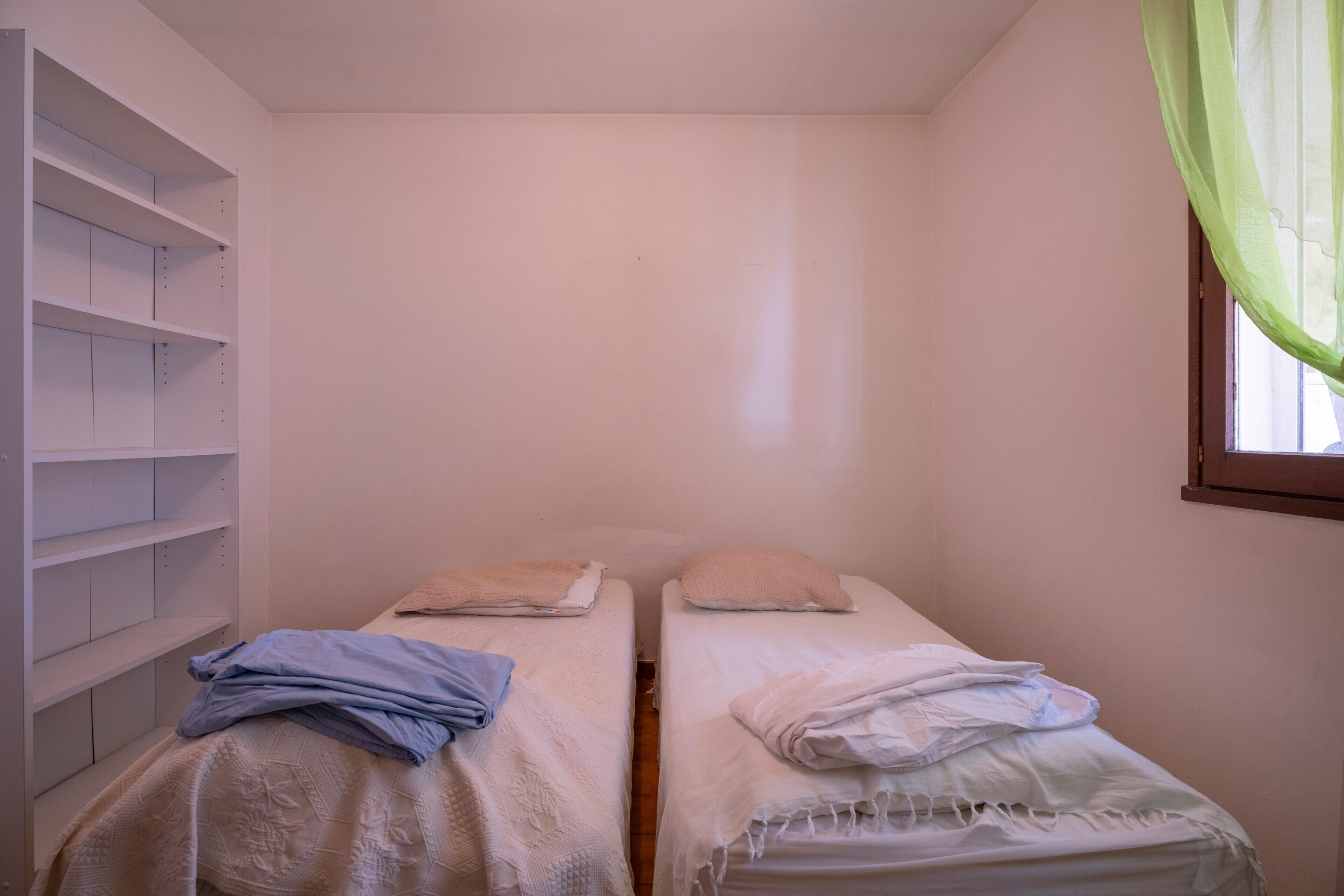 Ein kleines Zimmer mit zwei Betten, einem weißen Kleiderschrank und einem Fenster mit grünen Vorhängen.