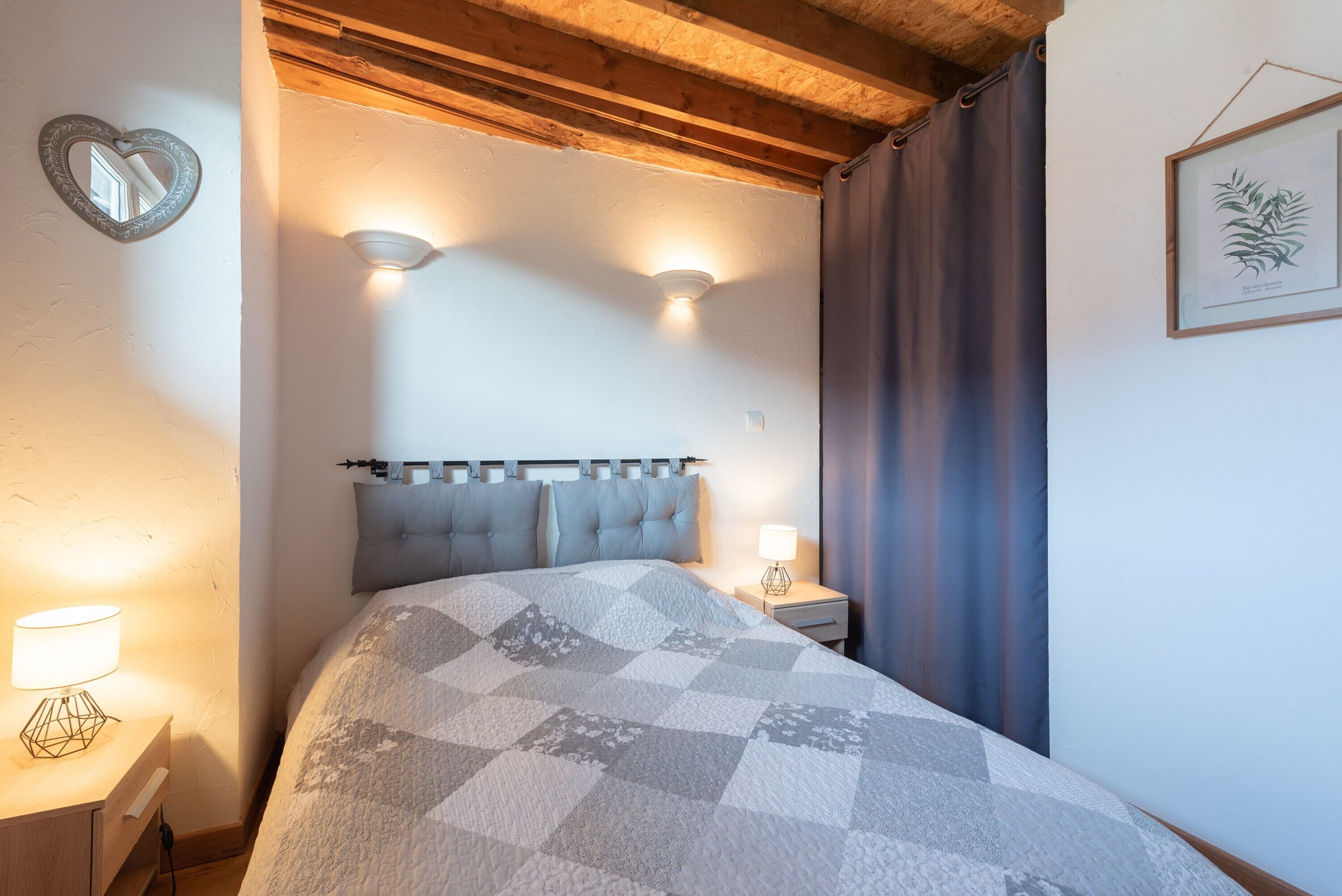 A bedroom with a bed, two bedside lamps, a headboard, a nightstand, a wall-mounted light, a heart-shaped mirror, and a framed botanical print.