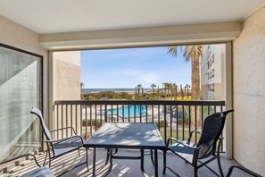 A balcony with a table and chairs, overlooking a pool and palm trees. - Surf & Racquet B122 - Hosted by Moore AI Rentals (Fernandina Beach)
