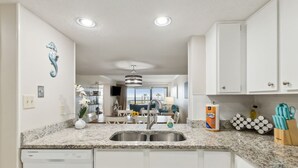 A modern kitchen with white cabinets, granite countertops, and stainless steel appliances. There is a dining area with a view of the ocean, and a hallway leading to a living space.