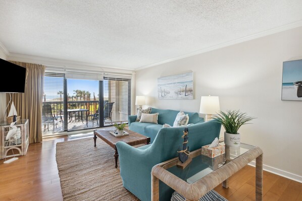 A living room with a teal sofa, a glass coffee table, and a view of the outdoors through large sliding glass doors. - Surf & Racquet B122 - Hosted by Moore AI Rentals (Fernandina Beach)