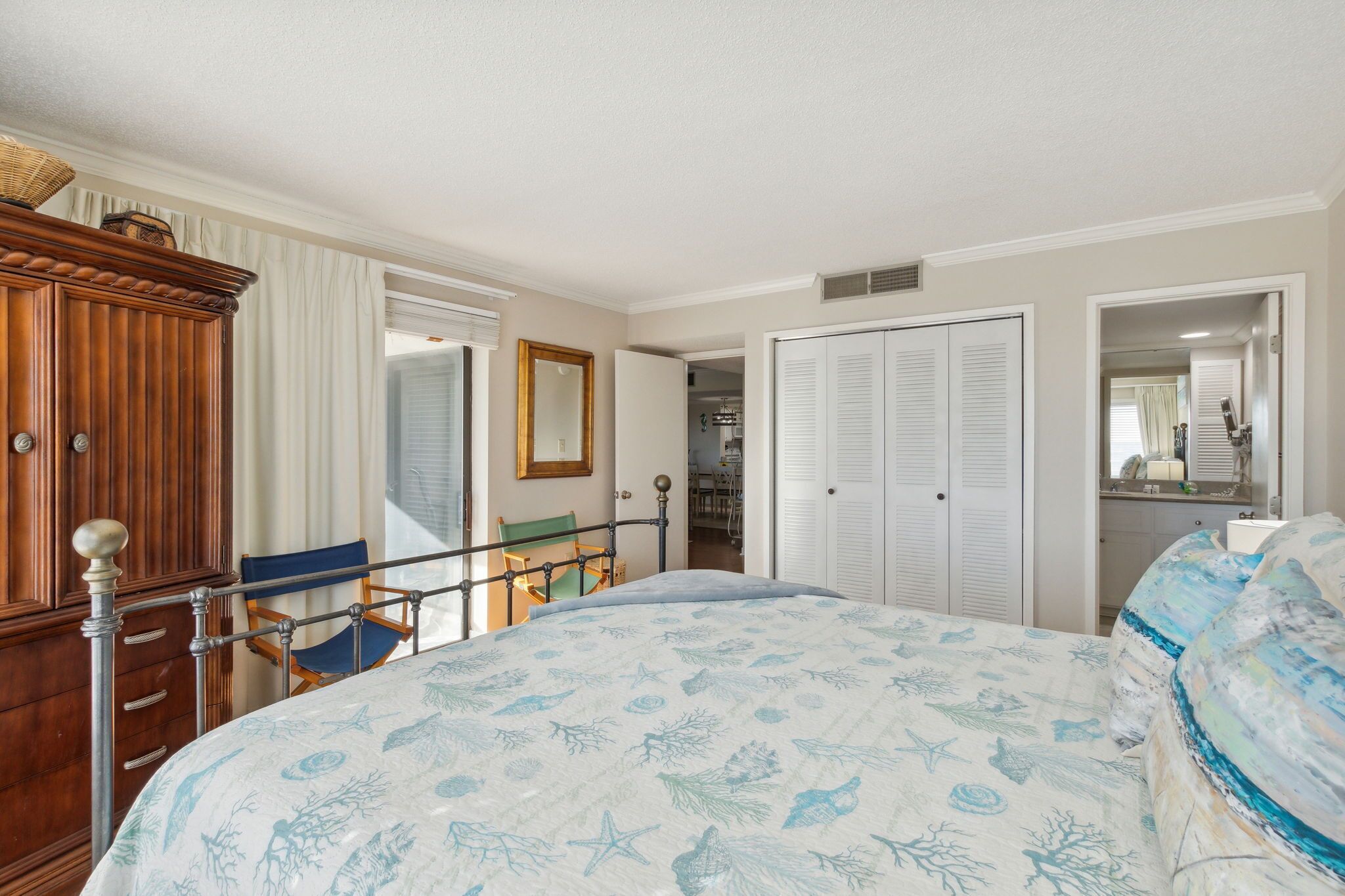 Un dormitorio con cama, un armario de madera, una silla, un espejo y una puerta que da a otra habitación.