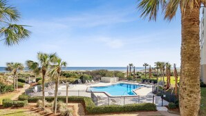 A swimming pool with a view of the ocean, surrounded by palm trees and a fenced area with lounge chairs.
