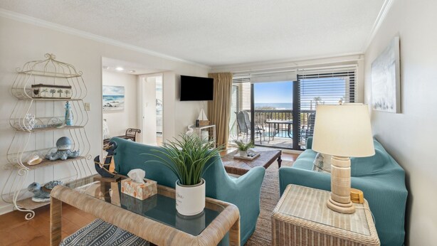A living room with teal sofas, a glass coffee table, a wicker side table, a metal shelf with decorative items, a flat-screen TV, and a balcony with a view.