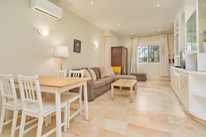 A living room with a sofa, a dining table with chairs, a small coffee table, a cabinet, and a window with curtains.