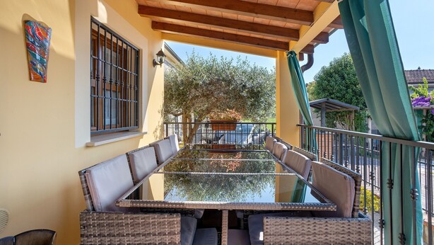 Un  área de estar al aire libre con techo, una mesa de vidrio y sillas de mimbre, vistas al jardín y un edificio con fachada amarilla.