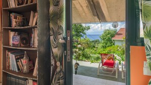 A room with a bookshelf, a red chair, and a view of greenery and a building. - Holiday Home 'Palati' with Sea View, Wi-Fi and Air Conditioning (Kefalonia)