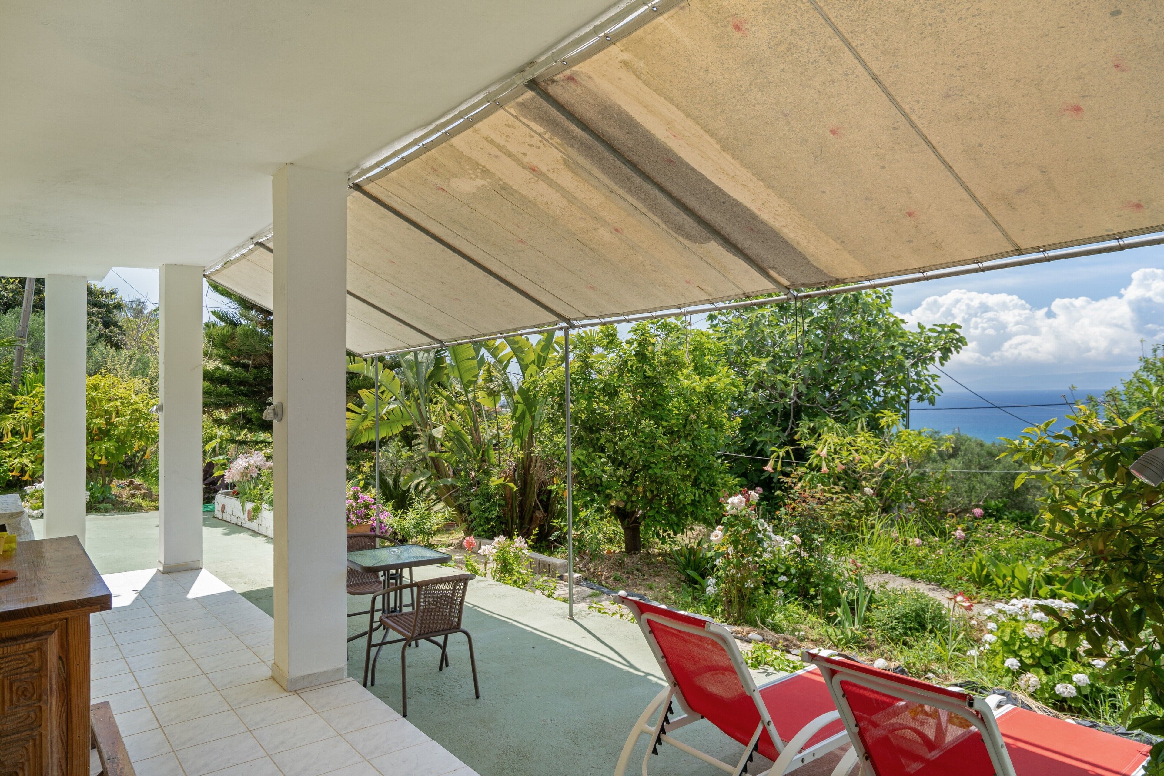 A covered patio with a table and chairs, a view of a garden, and a clear sky.