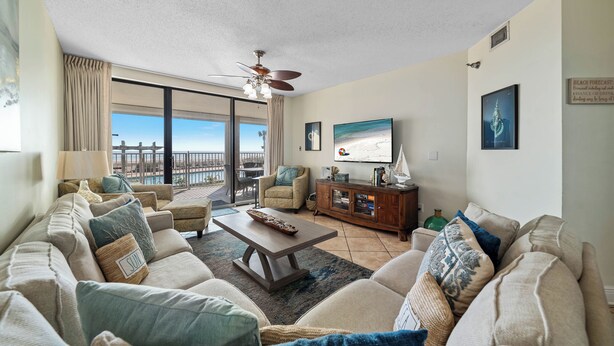 A spacious living room with a large sofa set, a coffee table, and a television. There is a balcony with a view of the ocean. - WHITE CAPS 105 - AFFORDABLE BEACHFRONT RELAXATION! (Orange Beach)