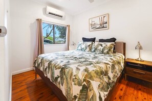 Une chambre à coucher comprenant un lit, une table de chevet, un appareil de climatisation, une fenêtre avec des rideaux et un tableau encadré accroché au mur.