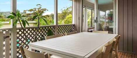 A covered outdoor area with a table and chairs, a wooden deck, and a view of greenery and trees.