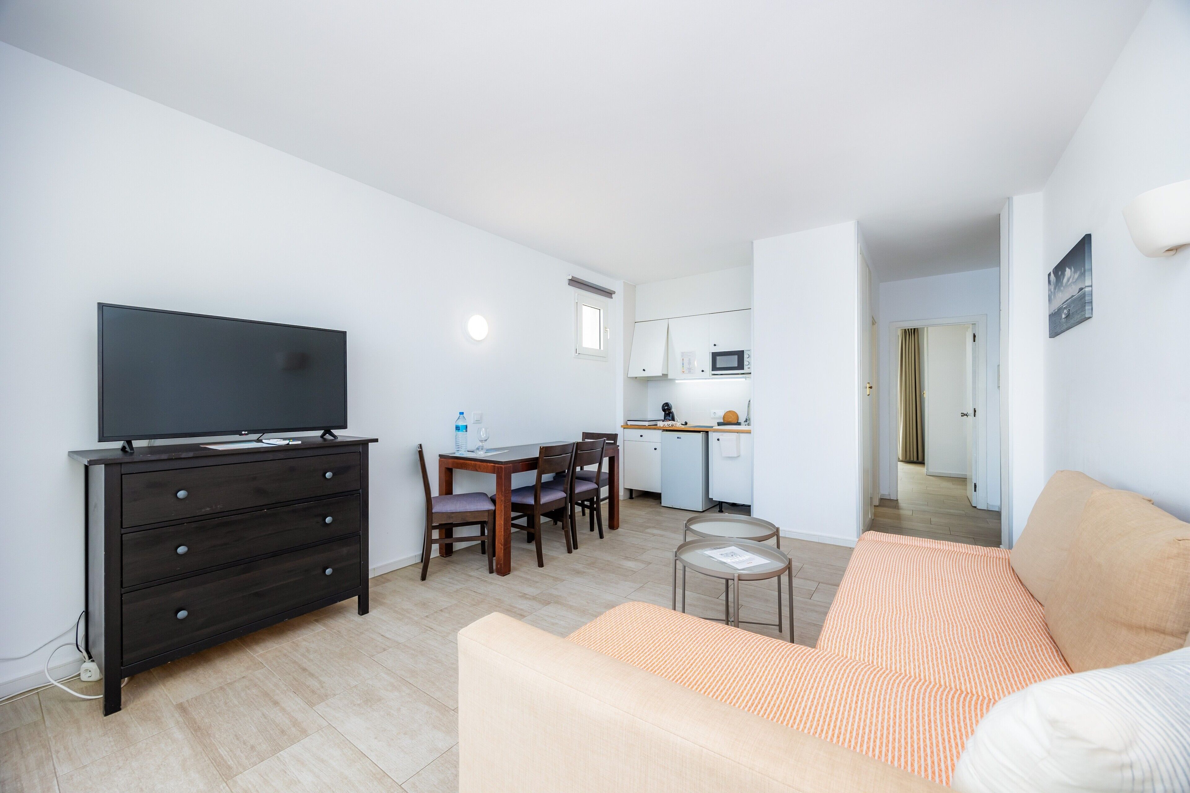 Un soggiorno moderno con una TV a schermo piatto, un comò in legno scuro, un divano beige, un'area pranzo con tavolo e sedie e una cucina sullo sfondo.