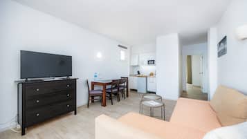 Un soggiorno moderno con una TV a schermo piatto, un comò in legno scuro, un divano beige, un'area pranzo con tavolo e sedie e una cucina sullo sfondo.