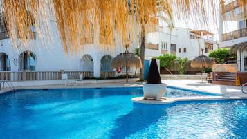 A swimming pool with a stone sculpture, white buildings with arched windows, and thatched umbrellas.