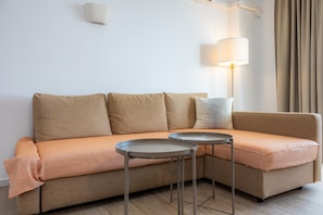 A beige sectional sofa with striped cushions, two small round tables, a wall-mounted lamp, and a window with curtains. - Apartment 'Sea View Room 106' with Shared Pool, Wi-Fi and Air Conditioning (Portocolom)