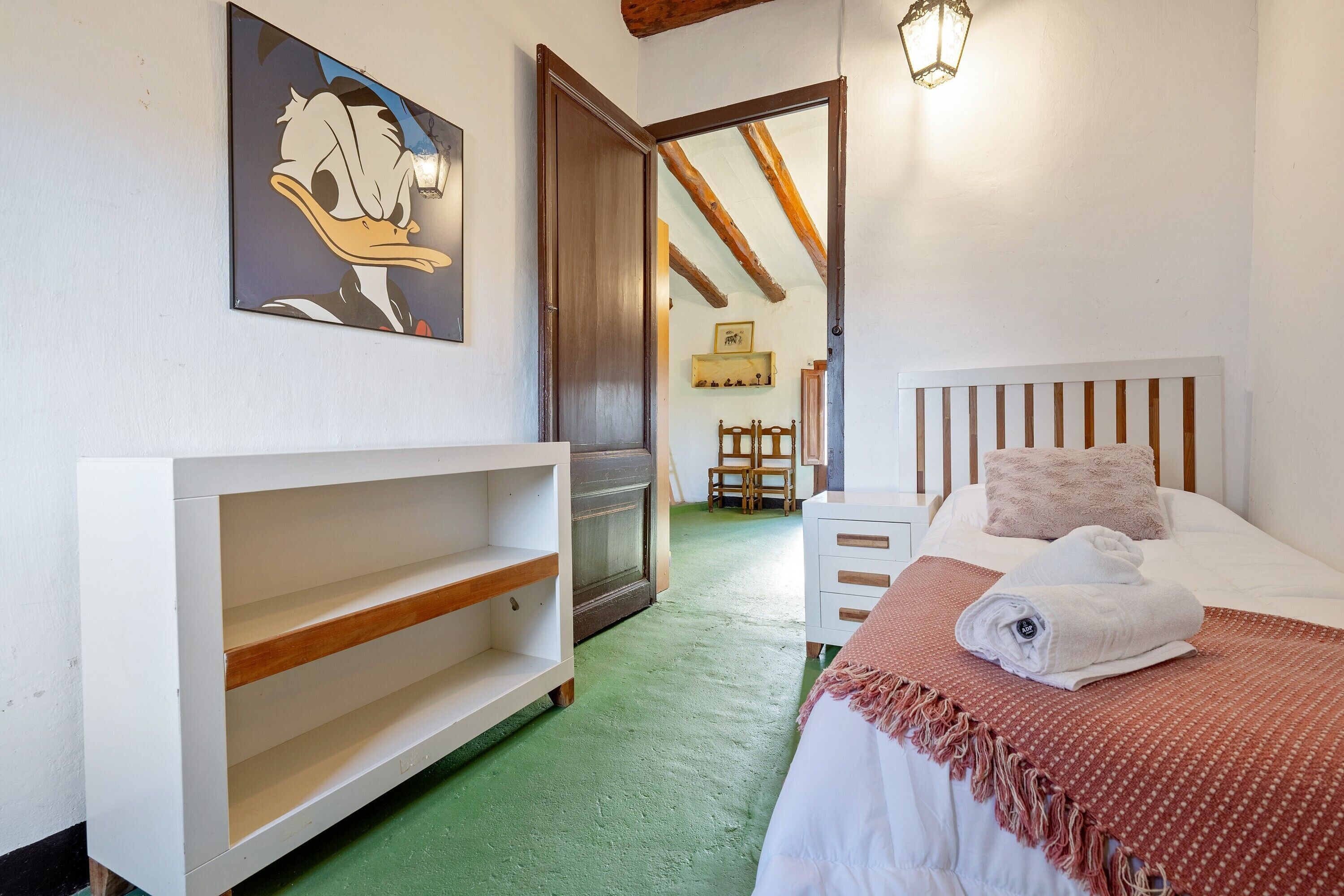 Ein Schlafzimmer mit einem Bett, einem weißen Nachttisch, einem Holzstuhl, einer Regaleinheit und einem Bild an der Wand.