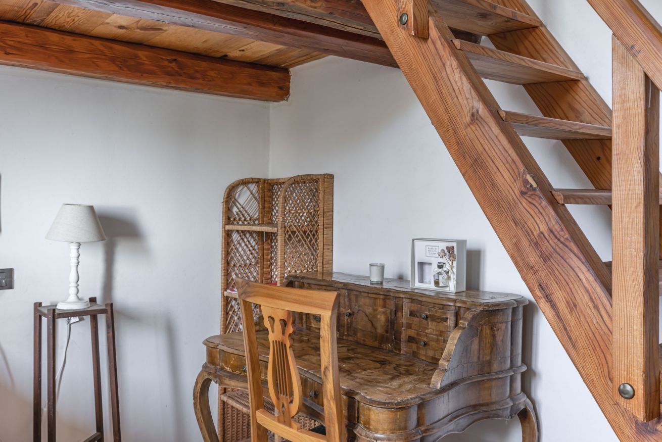 Una habitación con una escalera de madera, una silla de madera, una mesita con una lámpara y un sillón de mimbre.
