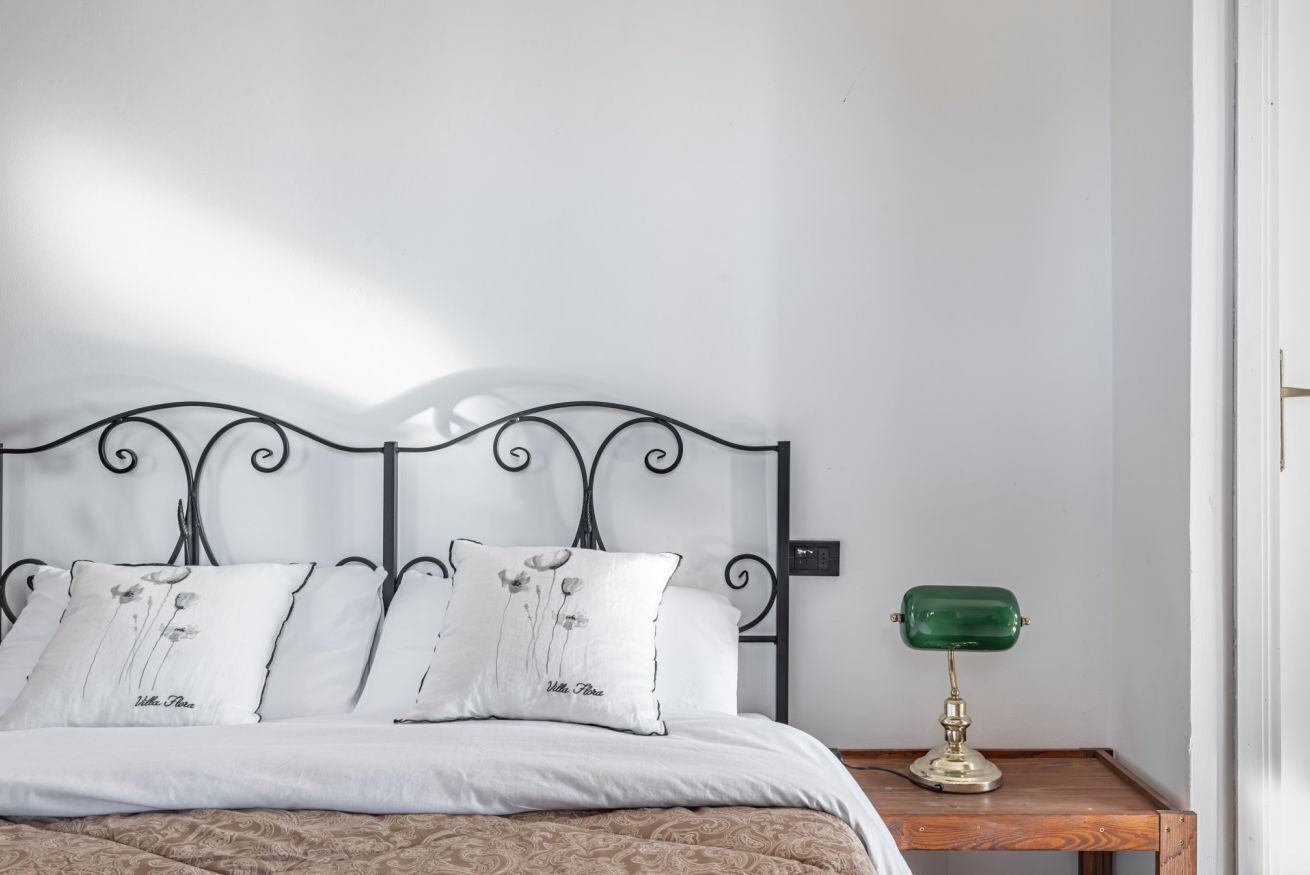 Uma cama com cabeceira de metal, roupa de cama branca e abajur verde.