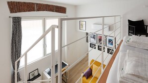 Una habitació amb un llit, una taula de treball, una finestra amb cortines, una escala i una paret amb quadres emmarcats.