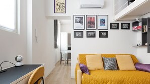 Habitació moderna amb escriptori, un sofà groc i una zona de menjador al fons.