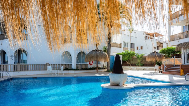 A swimming pool with a stone sculpture, white buildings with arched windows, and thatched umbrellas.