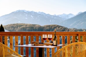 Une table en bois, avec deux verres de vin rouge, une bouteille et une assiette de nourriture, sur un balcon offrant une vue sur la montagne.