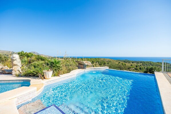A swimming pool with a view of the sea, surrounded by greenery and stone structures.