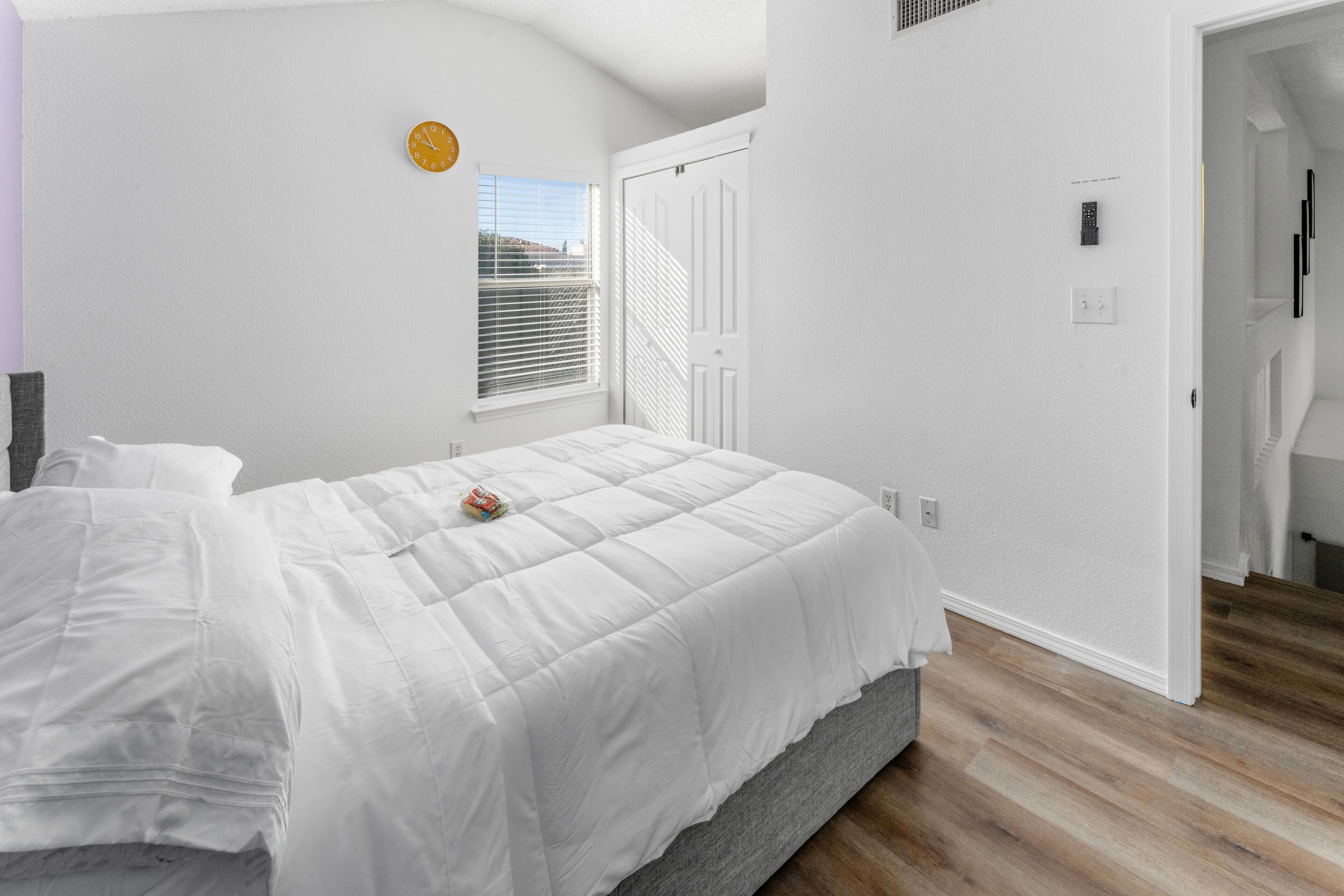 Un dormitorio con cama, ventana con persianas, una puerta y un reloj en la pared.