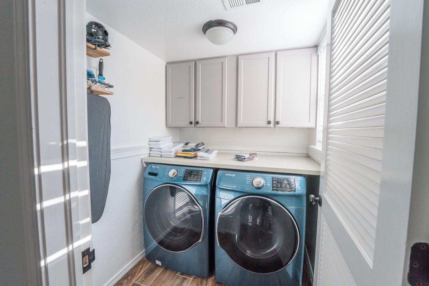 A compact laundry room with two front-loading washing machines, a countertop with various items, and white cabinets above.