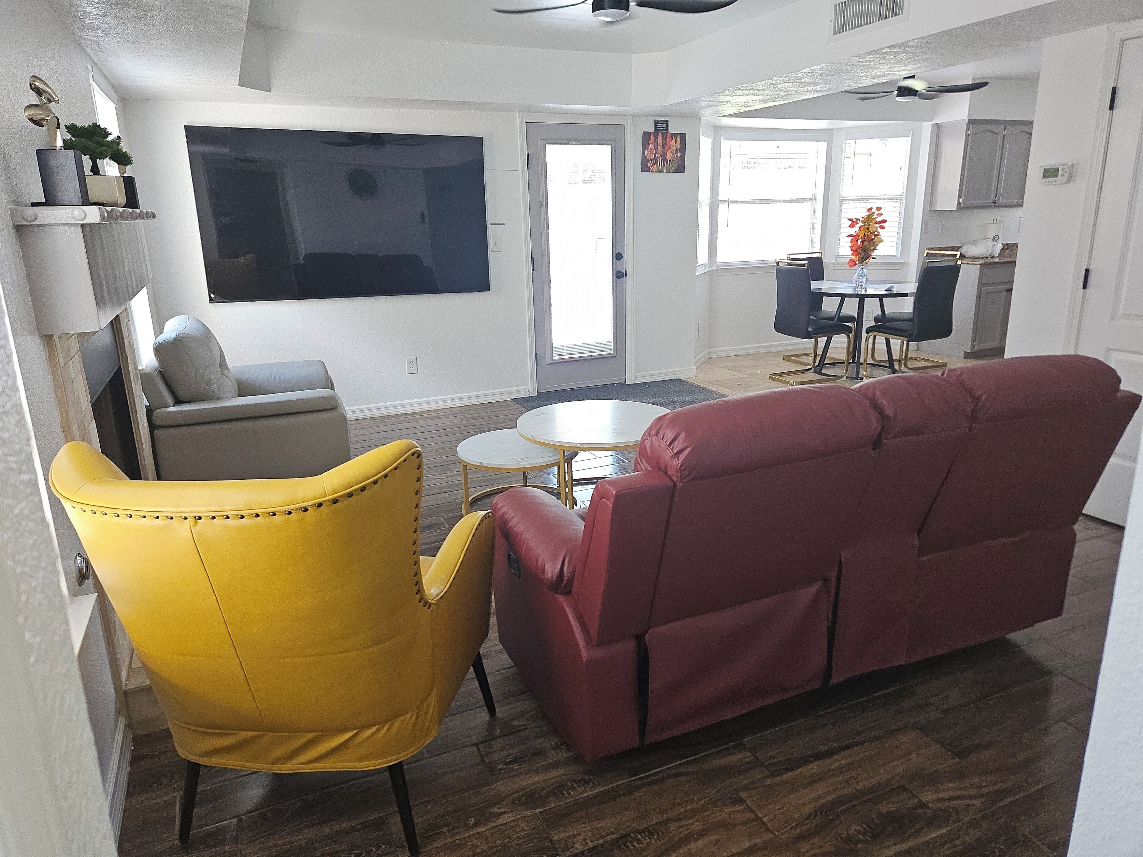 A living room with a yellow armchair, a red sofa, a flat-screen TV mounted on the wall, a dining area with a table and chairs, and a kitchen in the background.