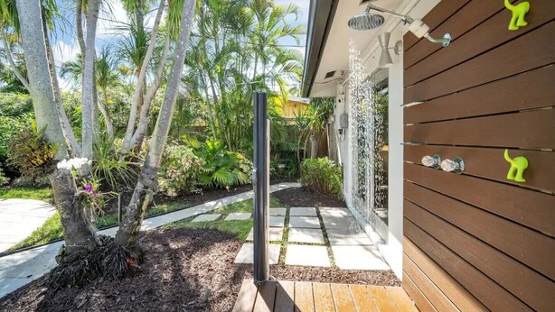 Pareti esterne in legno con zona doccia, un vialetto che conduce a una porta e un giardino con palme e piante.