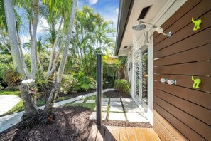 A wooden exterior wall with a shower area, a pathway leading to a door, and a garden with palm trees and plants.