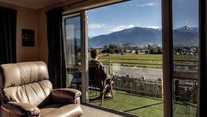 A room with a leather chair, a glass door leading outside, and a view of mountains and a golf course. - Close to Beach | Spacious | Outdoor Living - KK00379 (Kaikoura)