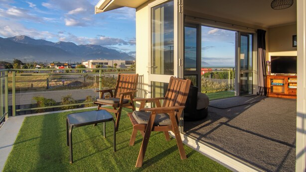 A balcony with a view of mountains, a seating area with chairs and a table, and a TV stand. - Close to Beach | Spacious | Outdoor Living - KK00379 (Kaikoura)