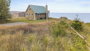 A single-story house with a green roof, a chimney, and a small porch. It is located near a body of water and surrounded by grass and shrubs.