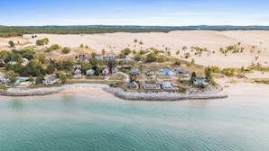 A coastal residential area with houses on stilts over water, surrounded by sand dunes and sparse vegetation.