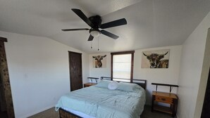 Une chambre à coucher comprenant un lit, un ventilateur de plafond, une tête de lit en bois, une table de chevet, une fenêtre avec des stores et deux tableaux représentant des taureaux, accrochés au mur.