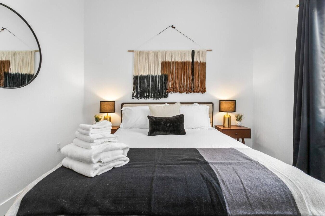 Een net opgemaakt bed met witte lakens, een zwarte plaid en een zwart kussen. Twee nachtkastjes met lampen, een wandspiegel en een decoratief macramé kunstwerk boven het bed.