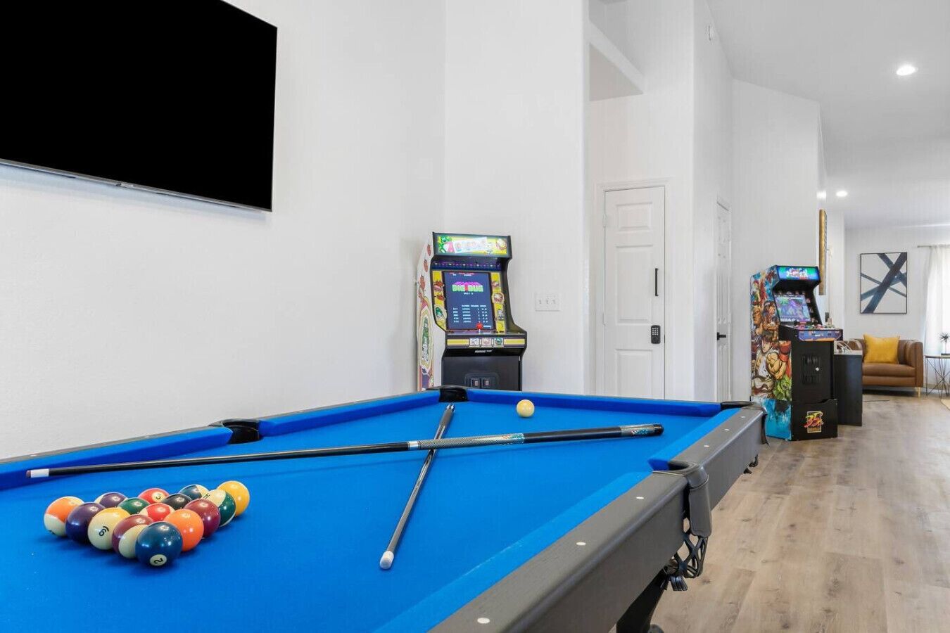 A pool table with balls and cues, two arcade machines, a sofa, and a TV mounted on the wall.